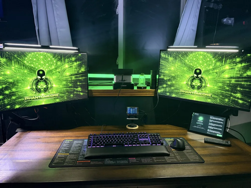 The build desk: two monitors, a keyboard and a PHNTM One running on the right, lit green